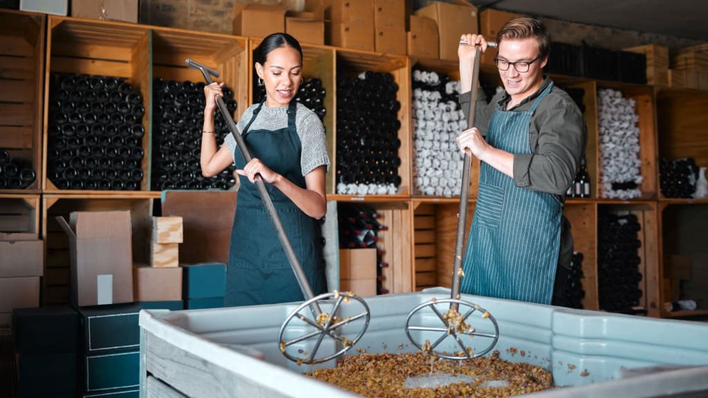 Two people in aprons, operating under a liquor manufacturer license, use metal tools to stir grape must in a large container; shelves of wine bottles and boxes can be seen in the background.
